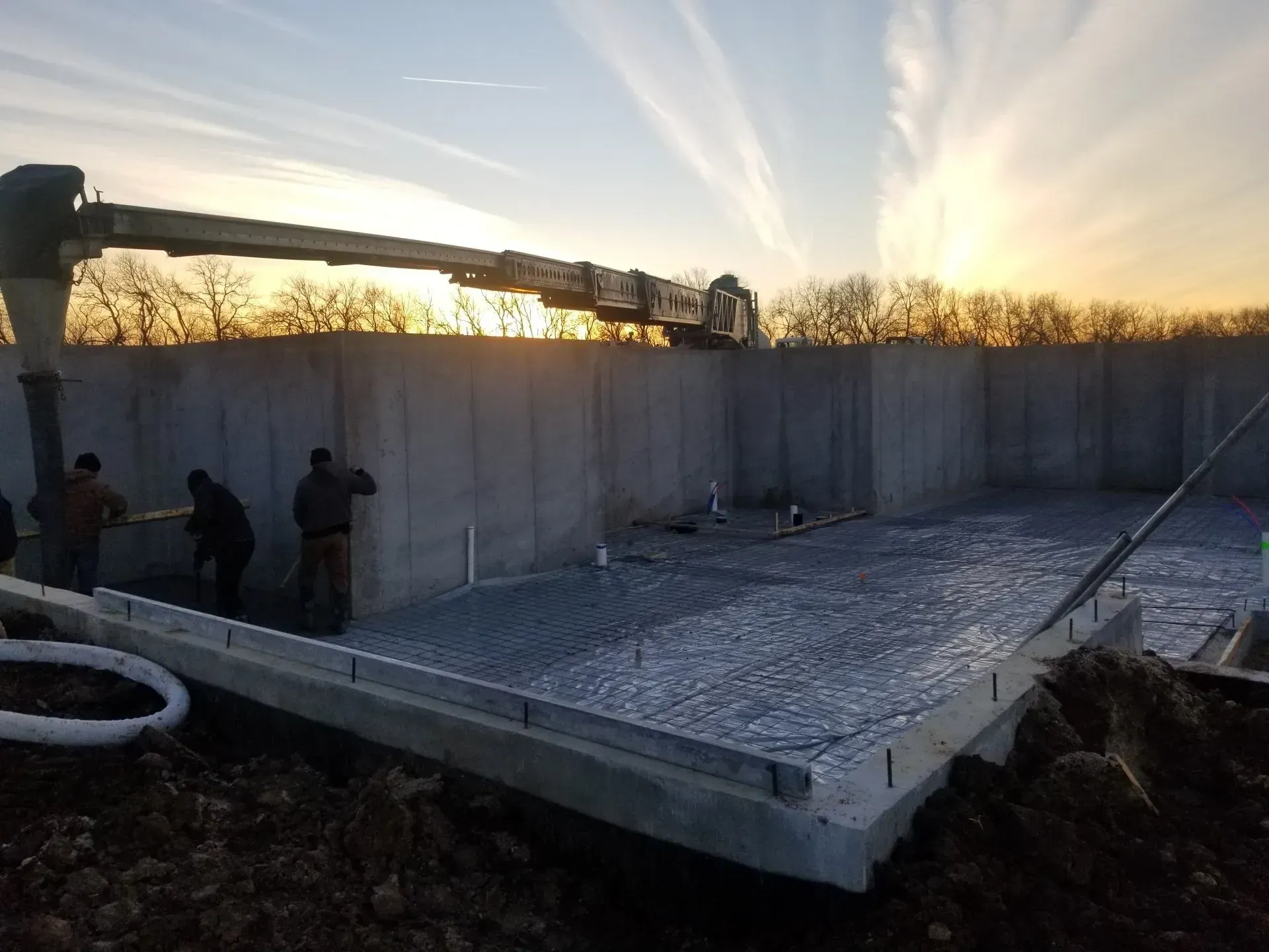 Concrete foundation being poured at sunset, with workers beside a partially built wall.