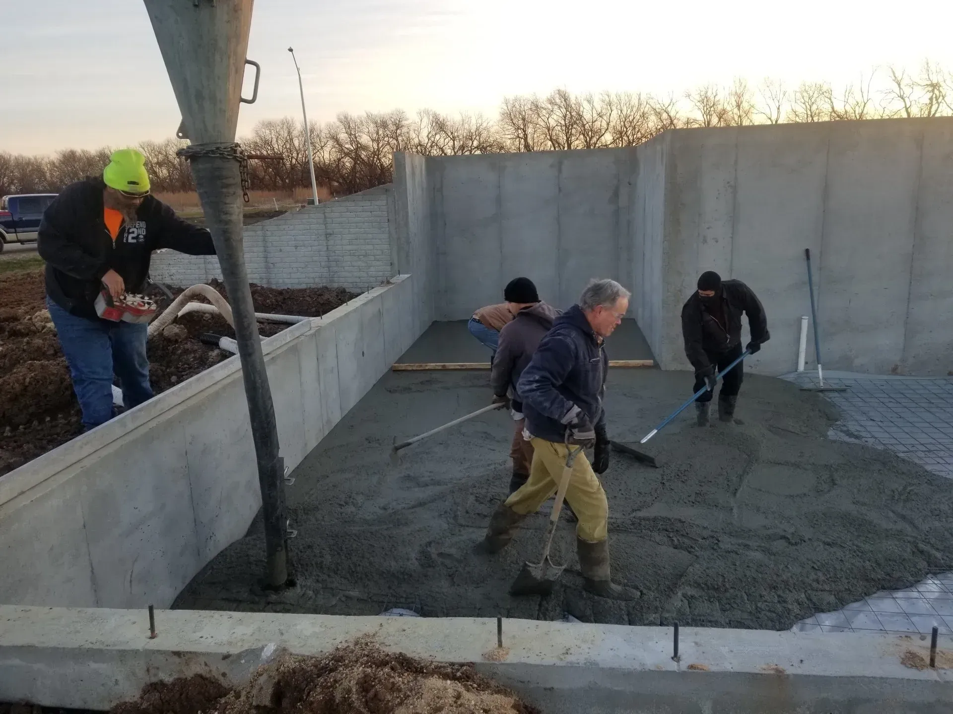 Workers smoothing wet concrete in a foundation area beside tall concrete walls at sunset
