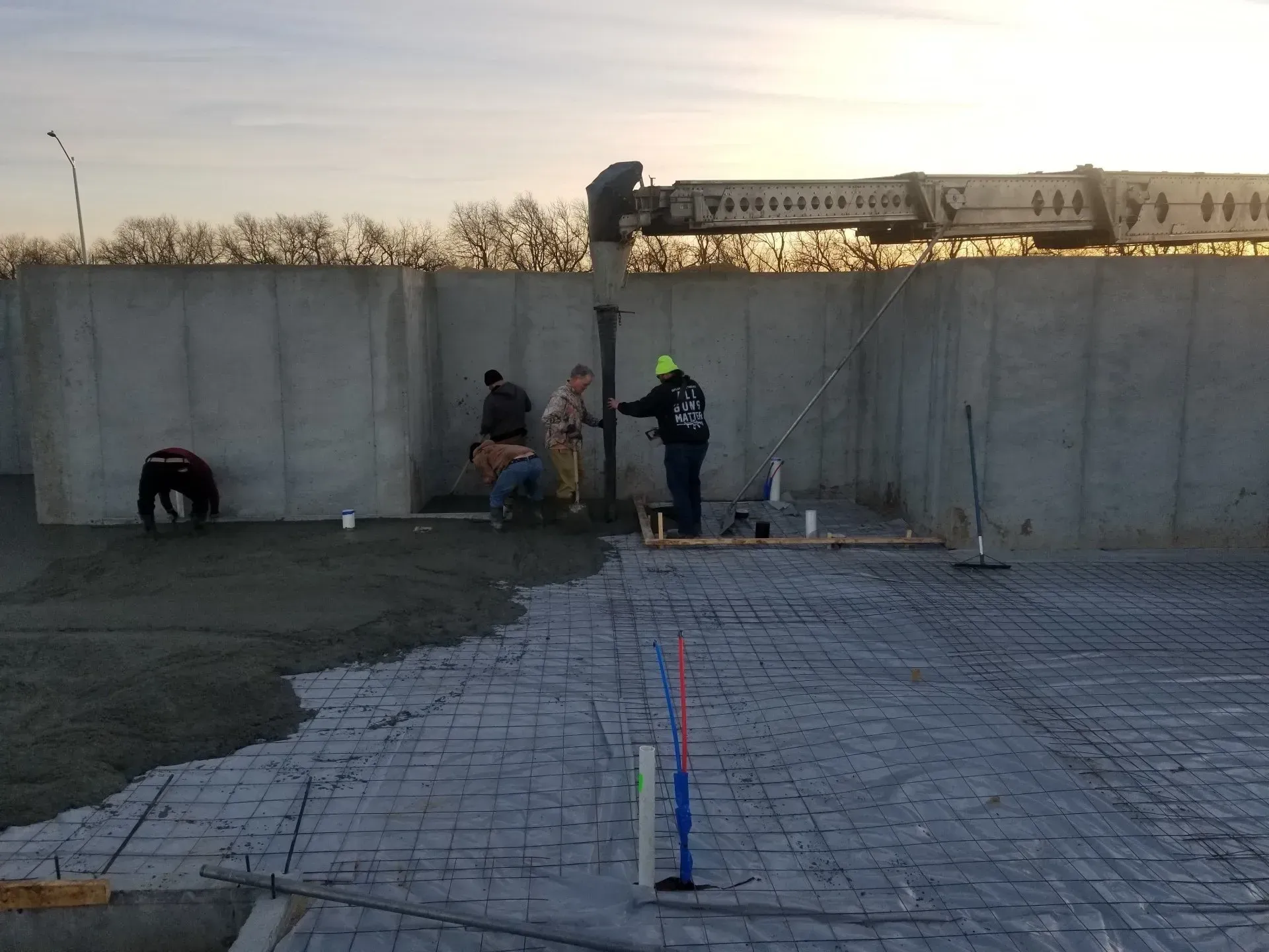 Construction workers by a concrete wall at dusk, with a paved path and survey markers in the foreground.