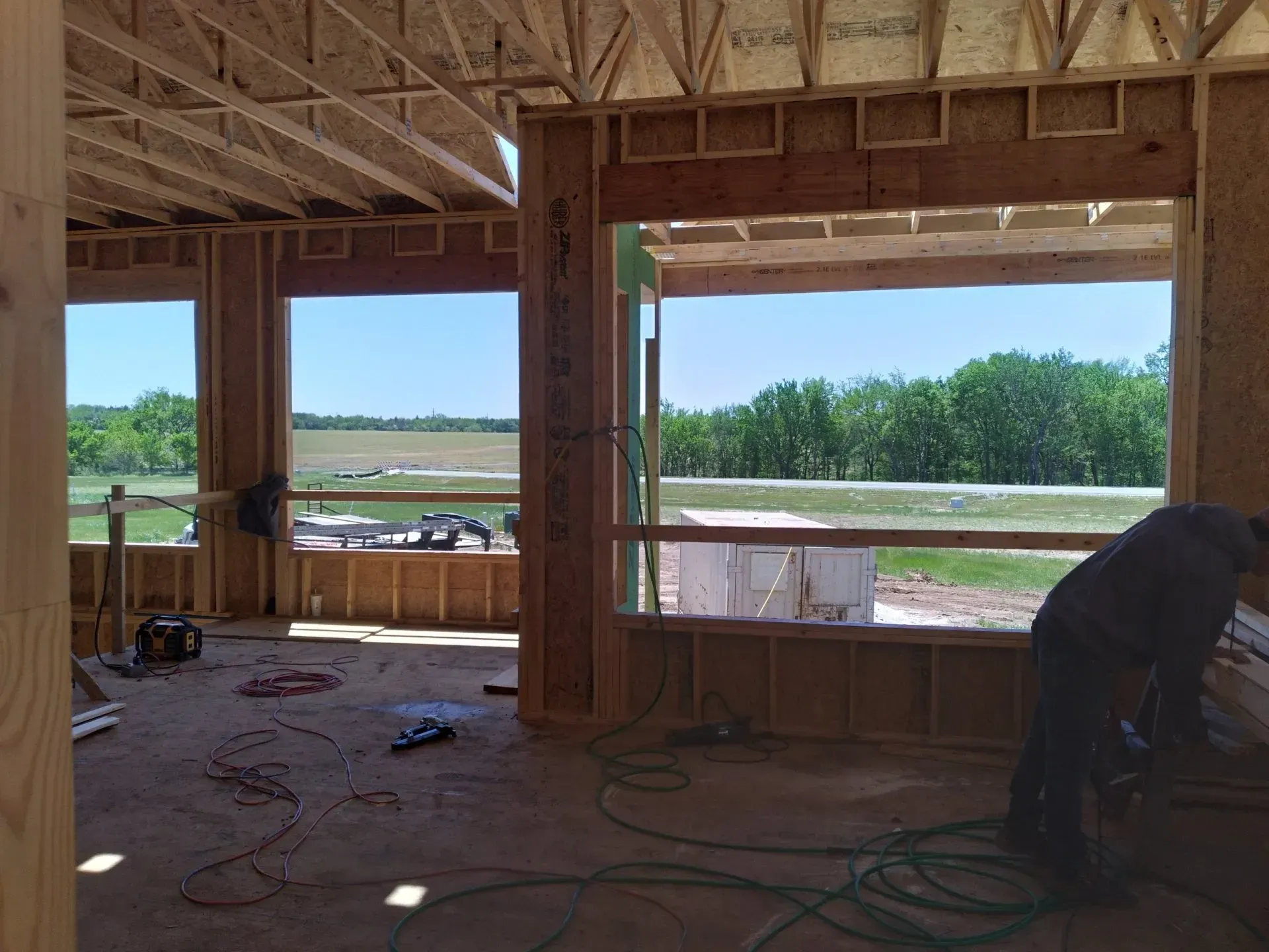 Interior of a wooden building under construction, with a worker on the right and fields visible through open framing.