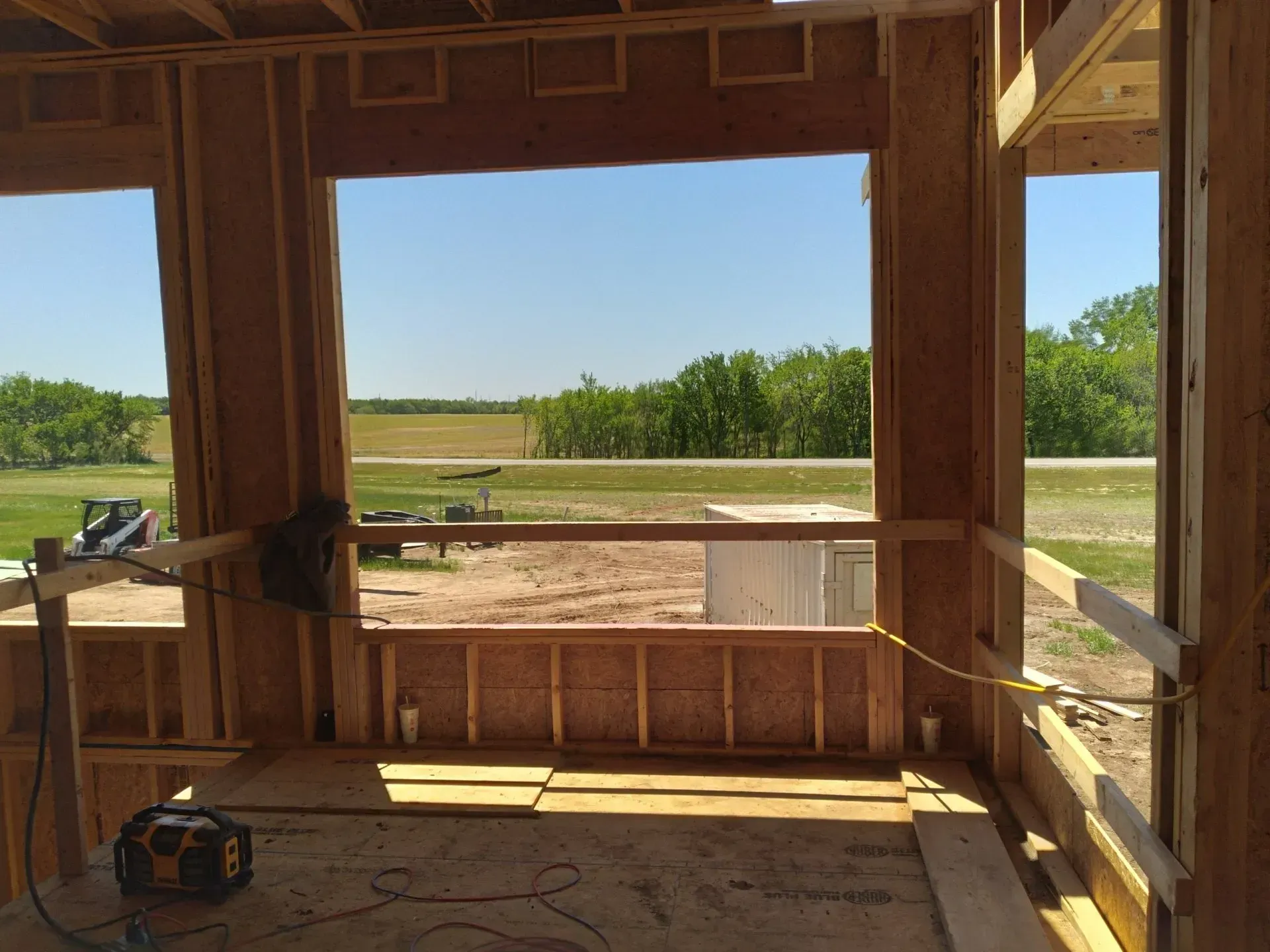 Inside an unfinished wooden cabin, looking out to a sunny field through open window and door frames.