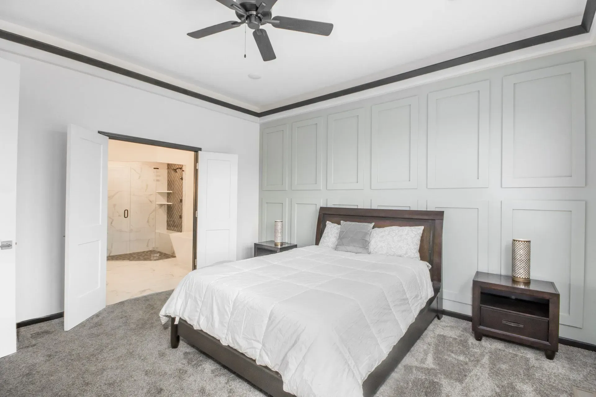 Bright bedroom with gray carpet, white paneled walls, bed, nightstand, ceiling fan, and open door to bathroom