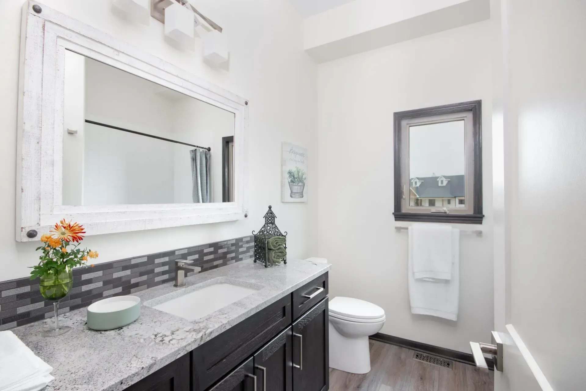 Bright white bathroom with dark vanity, large mirror, sink, toilet, and wall-mounted towel cabinet.
