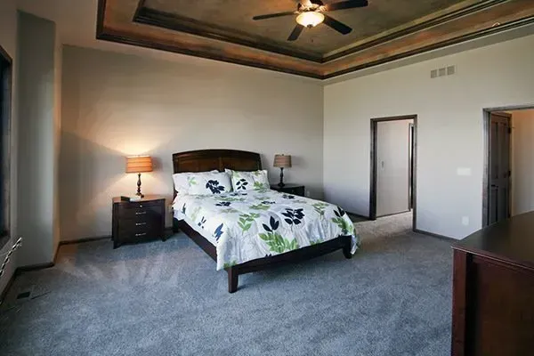 Spacious bedroom with a king bed, nightstand lamp, gray carpet, and ceiling fan.