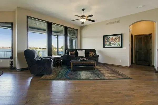 Spacious living room with large windows, dark armchairs, rug, ceiling fan, and hardwood floors.