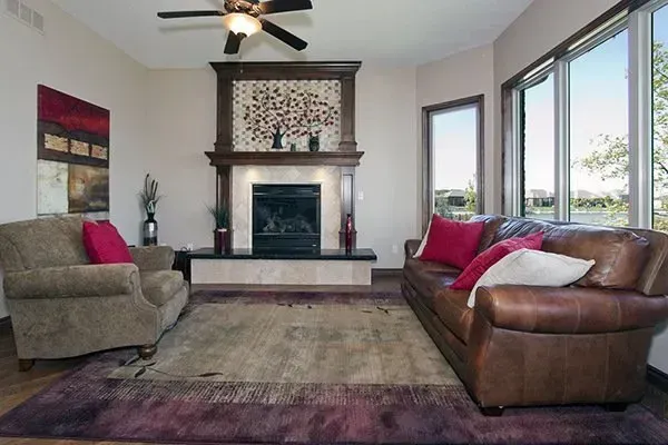Modern living room with stone fireplace, brown leather sofa, beige chair, and large windows.