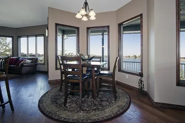 Dining area with round table on rug, dark wood floors, and large windows overlooking water.