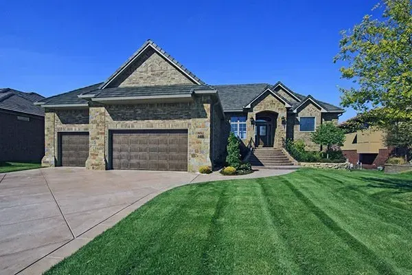 Suburban stone house with three-car garage, wide driveway, and neatly mowed green lawn under a blue sky