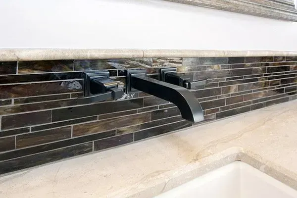 Black wall-mounted faucet over a beige sink and white tile backsplash.