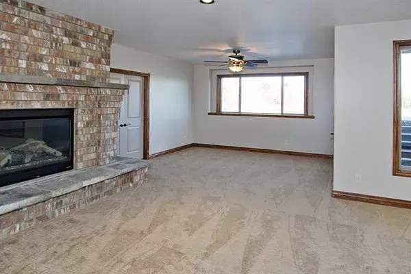 Empty living room with stone fireplace, beige carpet, and large windows across the far wall