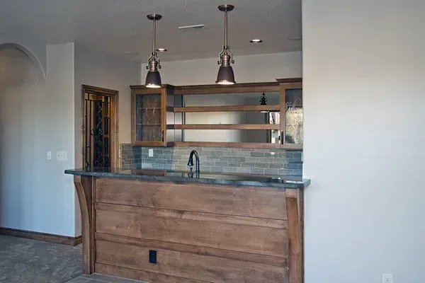 Empty home bar with wood cabinetry, granite counter, open shelves, and pendant lights against a white wall.