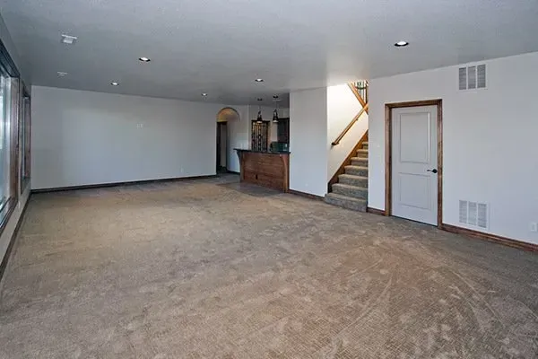 Empty carpeted basement room with gray walls, wood cabinet, and stairs by a closed door