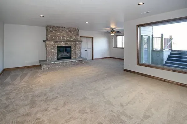 Spacious beige living room with stone fireplace, carpeted floor, and large window overlooking a patio