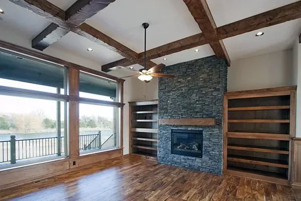 Empty living room with stone fireplace, wood floors, exposed beams, and large windows.
