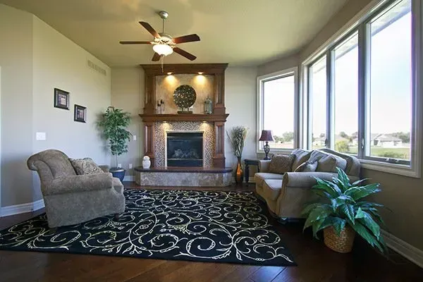 Bright living room with fireplace, patterned rug, gray seating, and large windows overlooking greenery