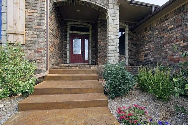 Front stone steps leading to a red front door under a covered entryway, with shrubs and flower beds nearby