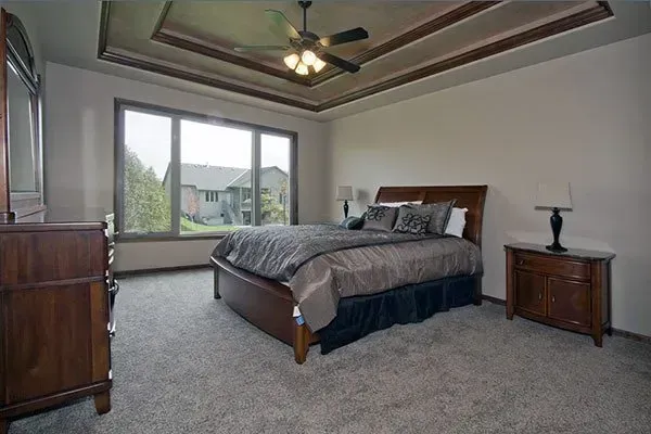 Spacious bedroom with a large bed, carpeted floor, ceiling fan, and a window overlooking greenery.