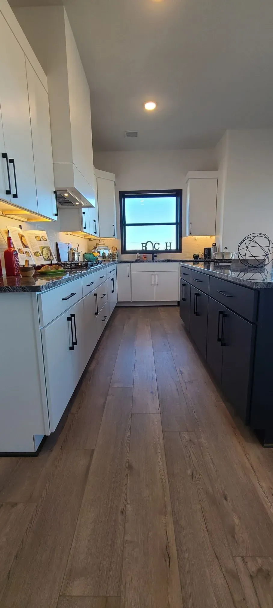 Narrow modern galley kitchen with white cabinets, wood floors, and a window at the far end.