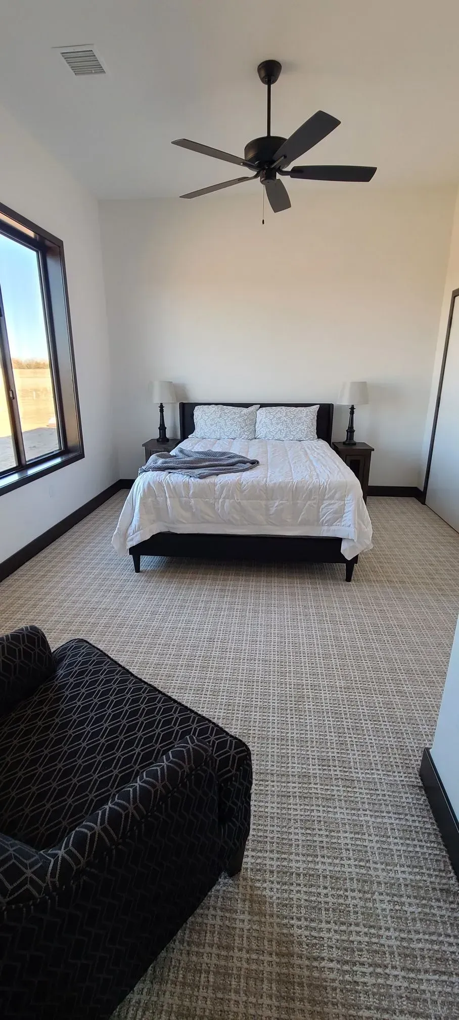 Bedroom with a bed, ceiling fan, large window, and black chair on a patterned rug.