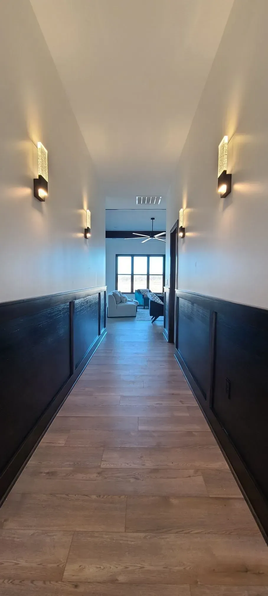 Narrow hallway with wood floors, dark wainscoting, wall sconces, and glass doors at the end.