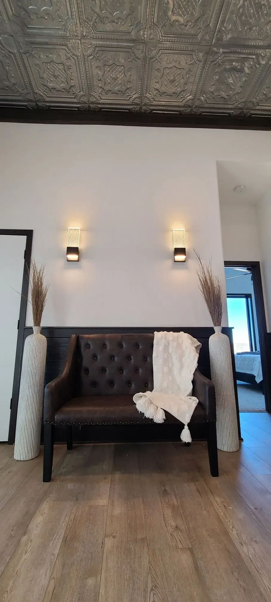 Black tufted loveseat with white throw in a hallway, under two wall sconces and a patterned ceiling.