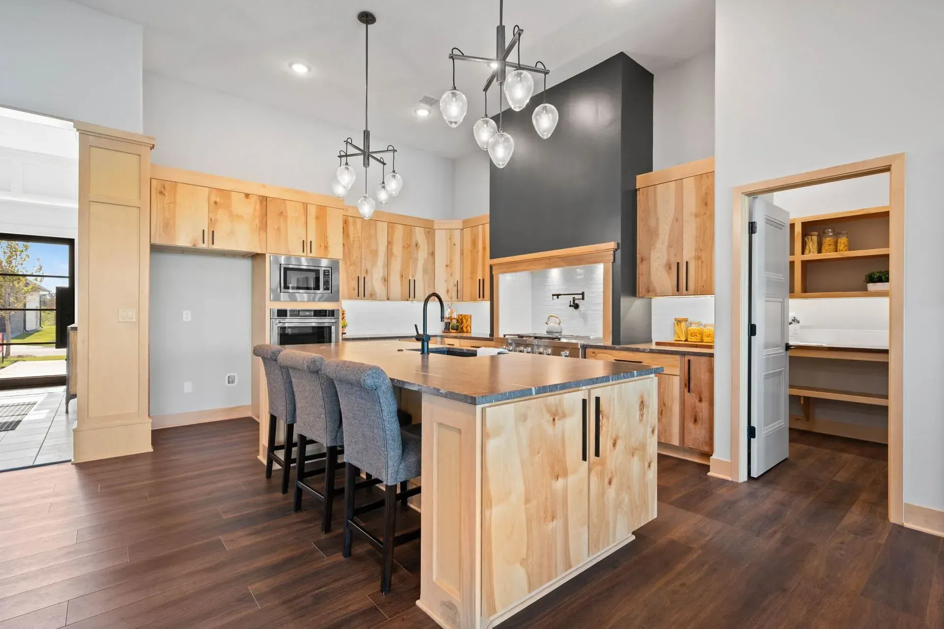 Modern kitchen with light wood cabinets, dark island stools, and pendant lights over dark flooring