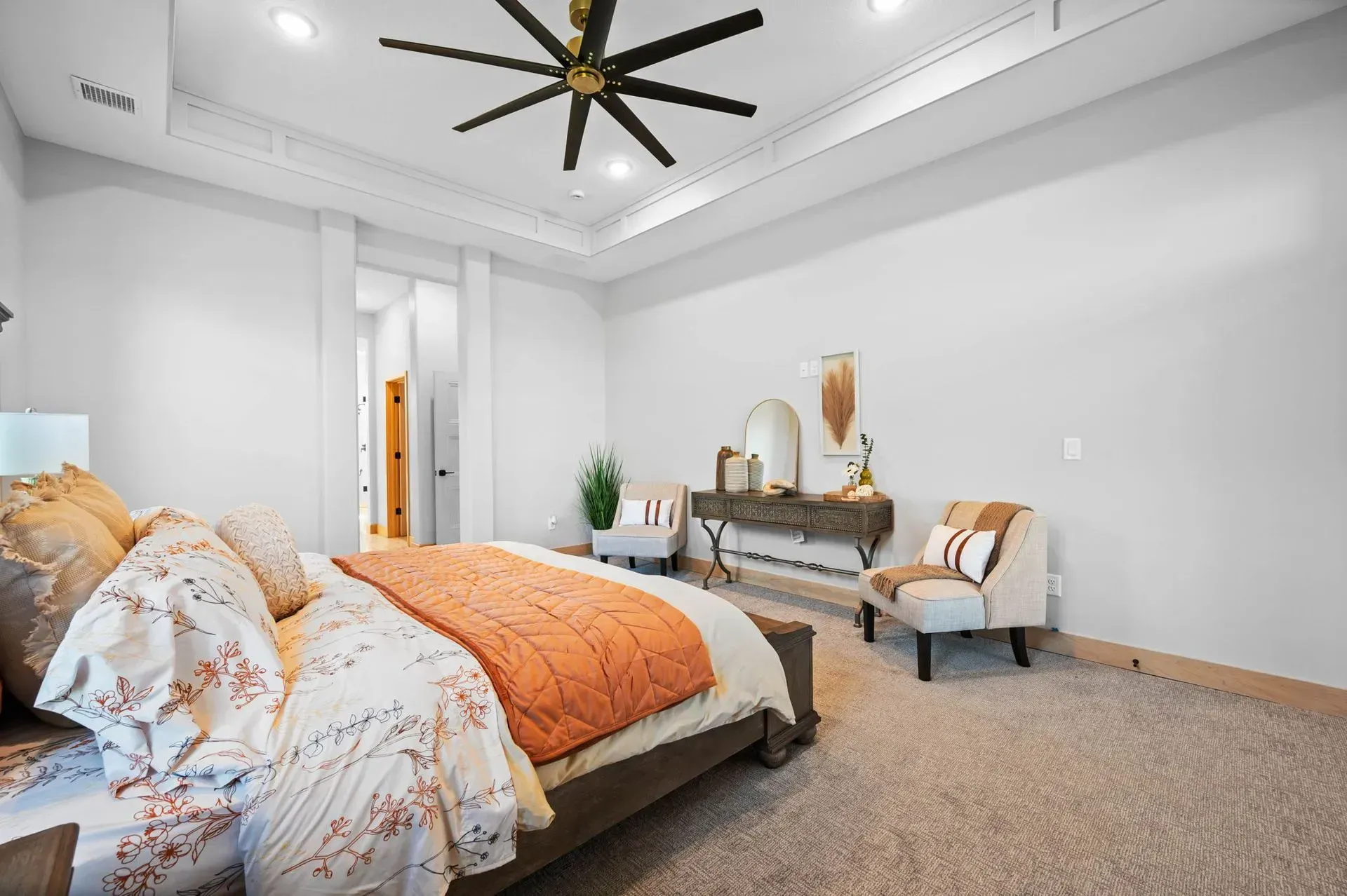 Bright bedroom with a large bed, orange accent blanket, ceiling fan, and vanity desk by the wall.