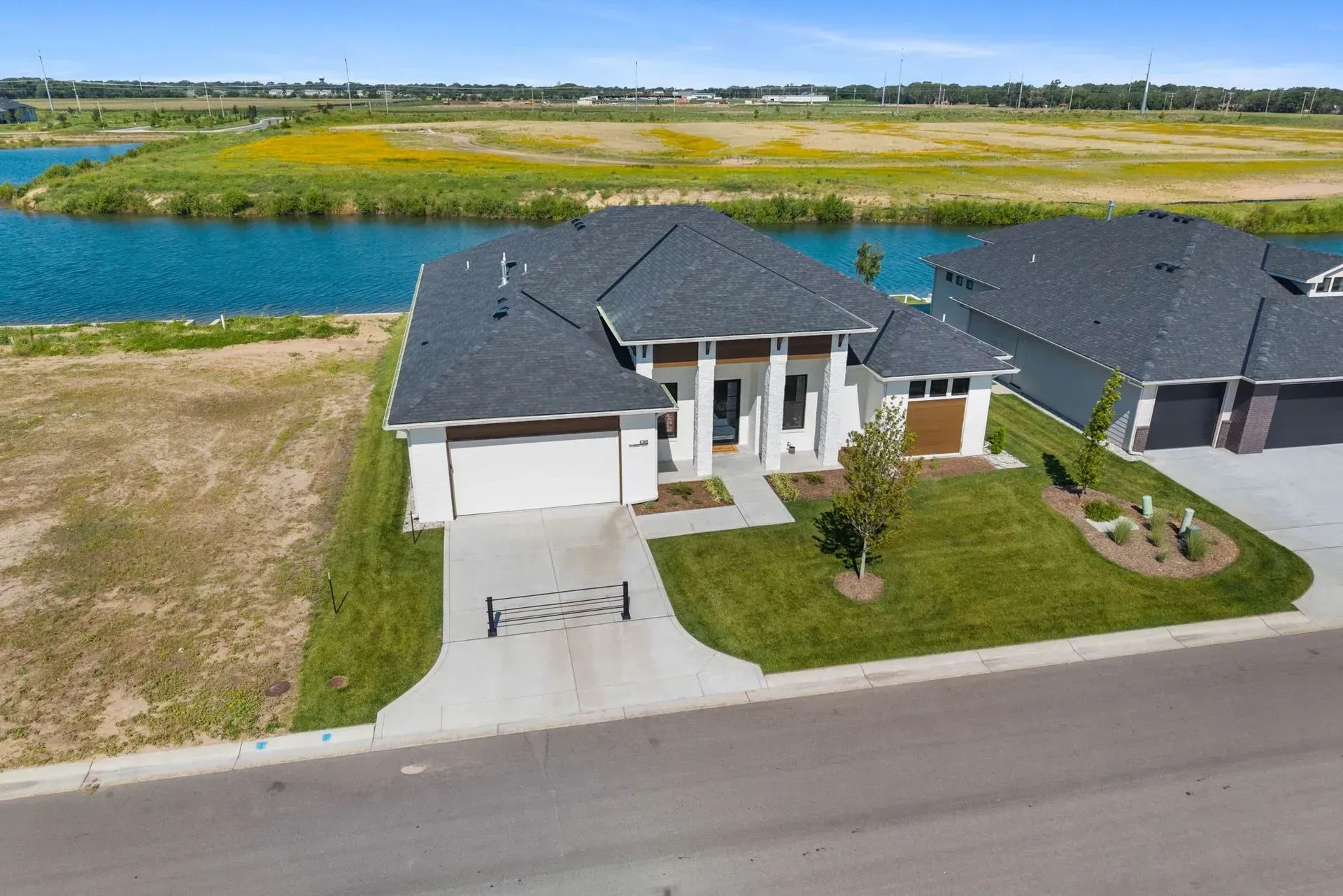 Aerial view of a modern house beside a road, with a driveway, lawn, and a lake behind it.