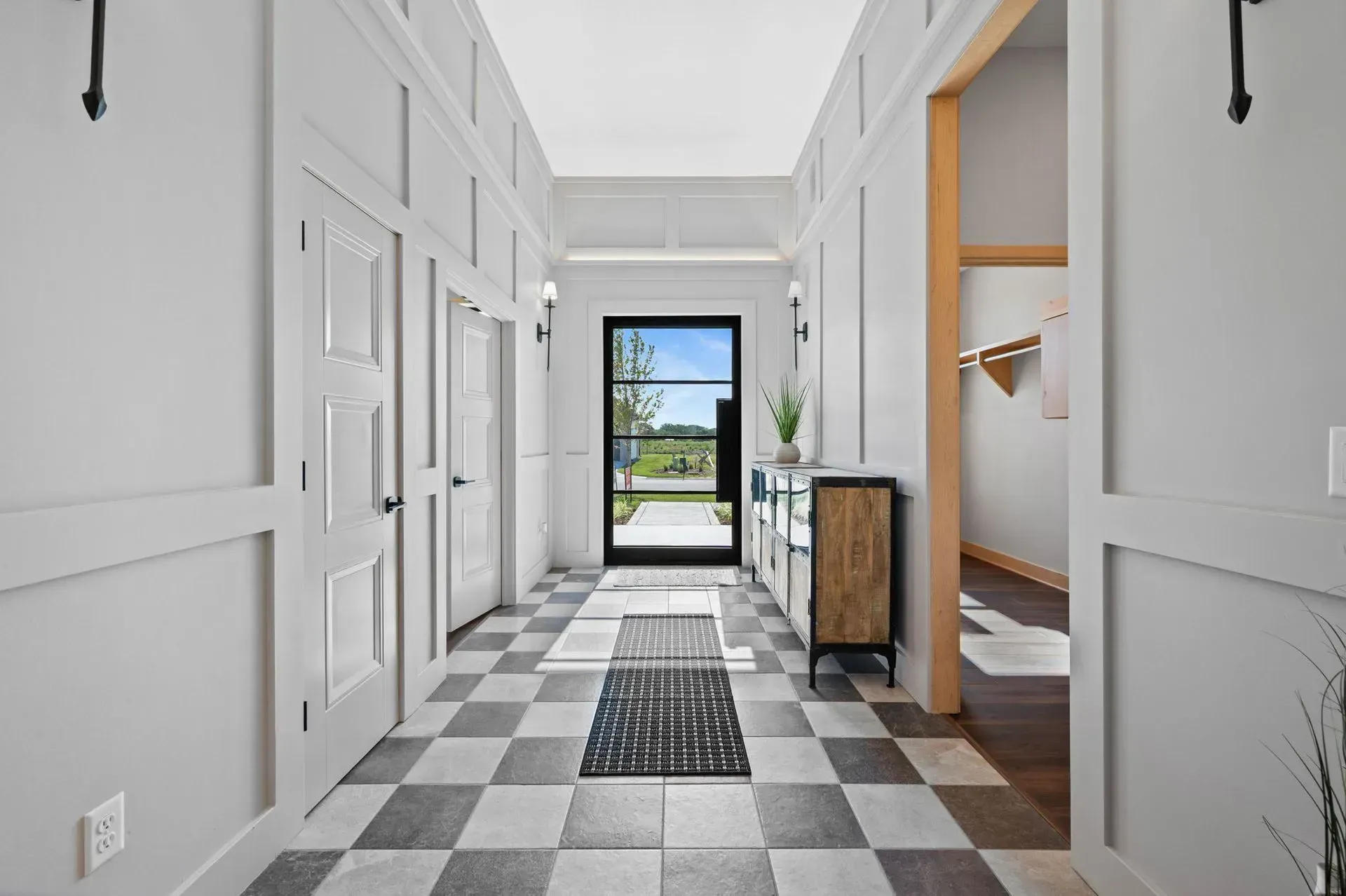 Modern hallway with black-and-white checkerboard floor, white paneled walls, and sunlight through a glass door.