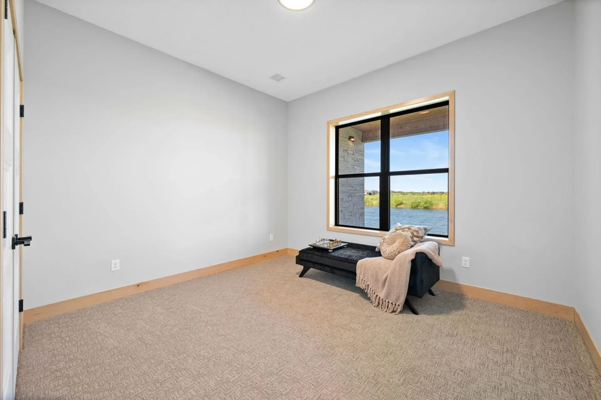 Empty carpeted bedroom with white walls, a window view, and a black bench with a throw blanket