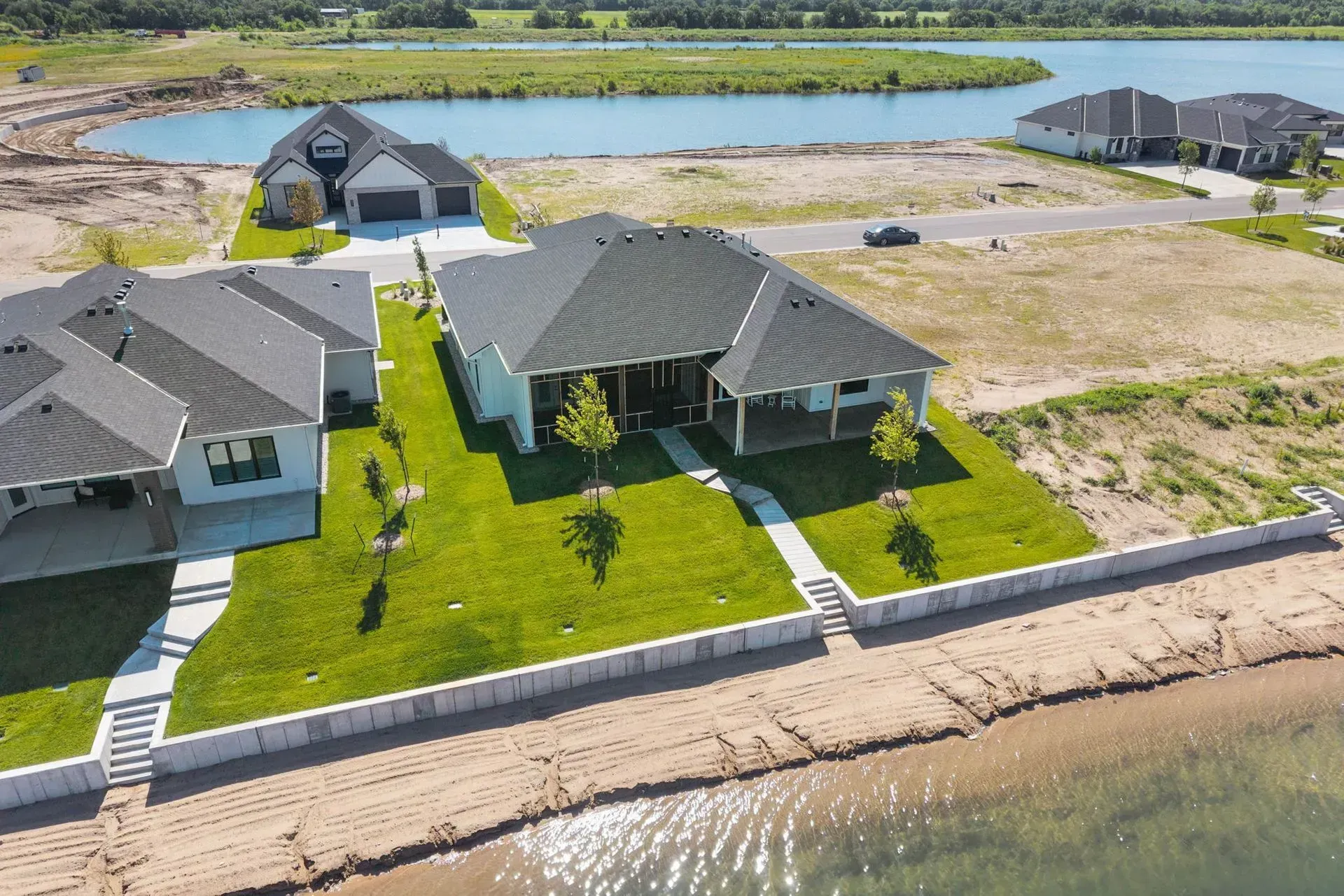 Aerial view of waterfront homes with green lawns, a seawall, and a sandy shoreline.