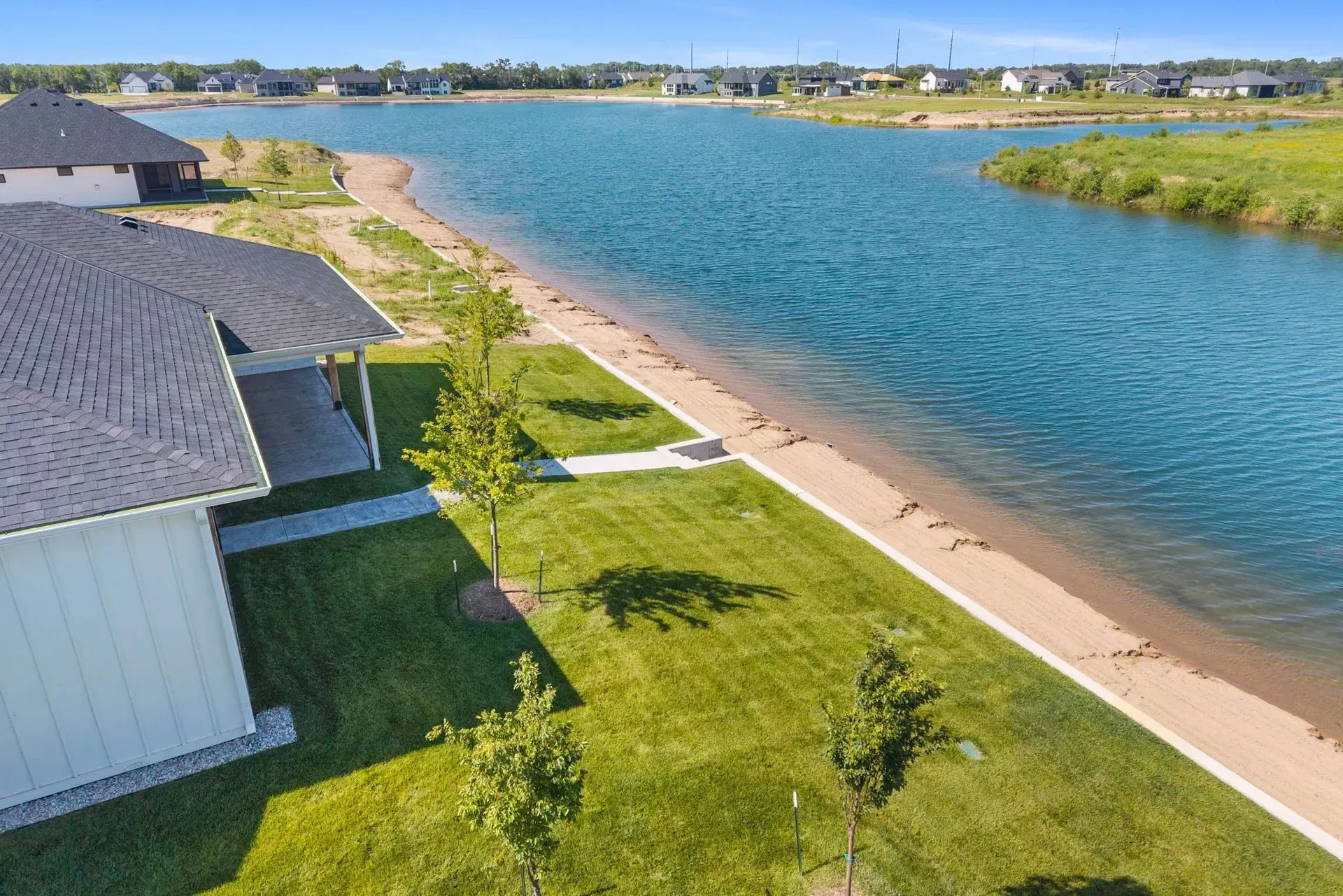 Aerial view of a waterfront home with green lawn, sandy shore, and blue canal under clear skies
