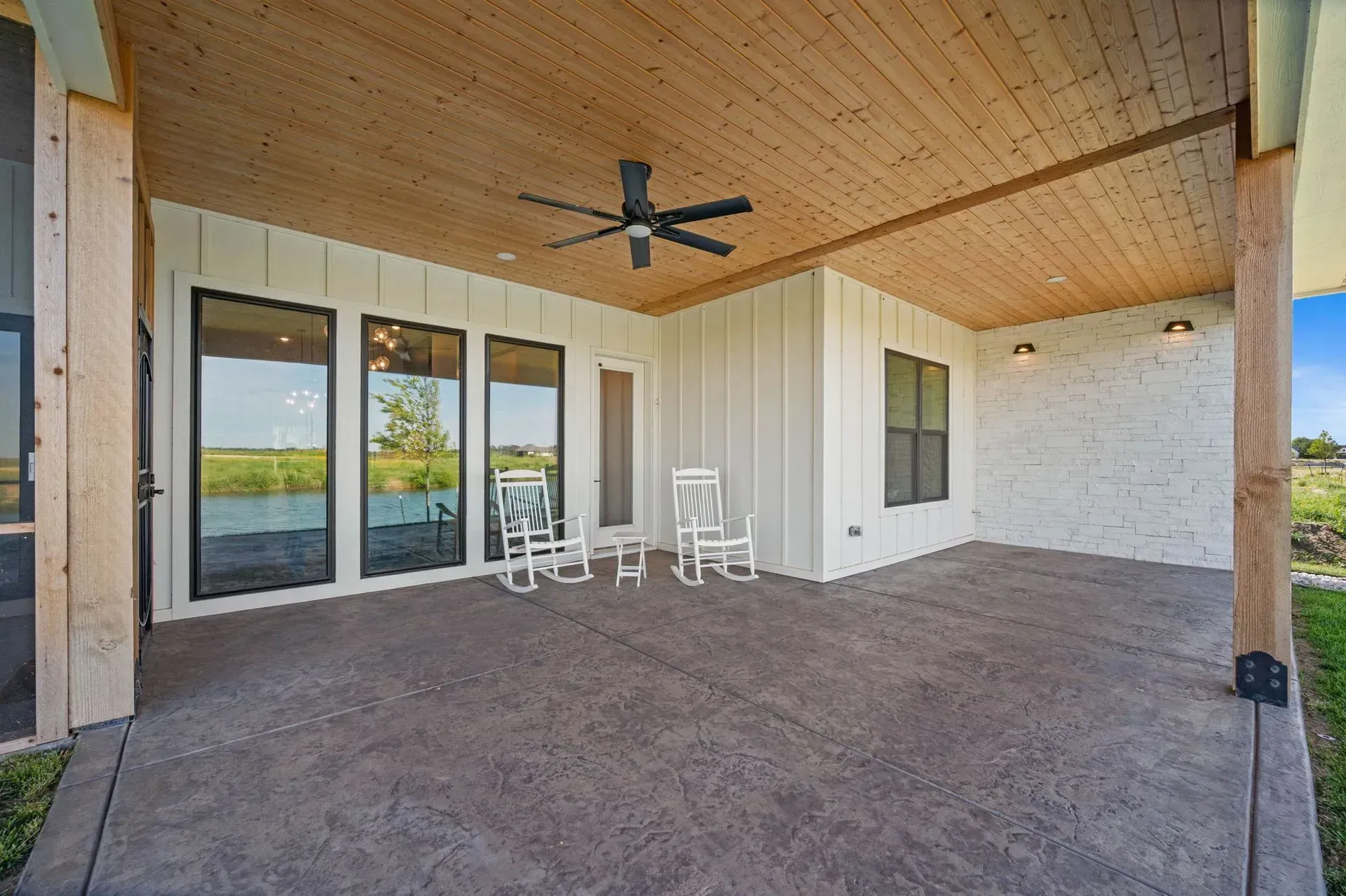 Covered patio with wood ceiling, ceiling fan, glass doors, and two white chairs overlooking a grassy yard
