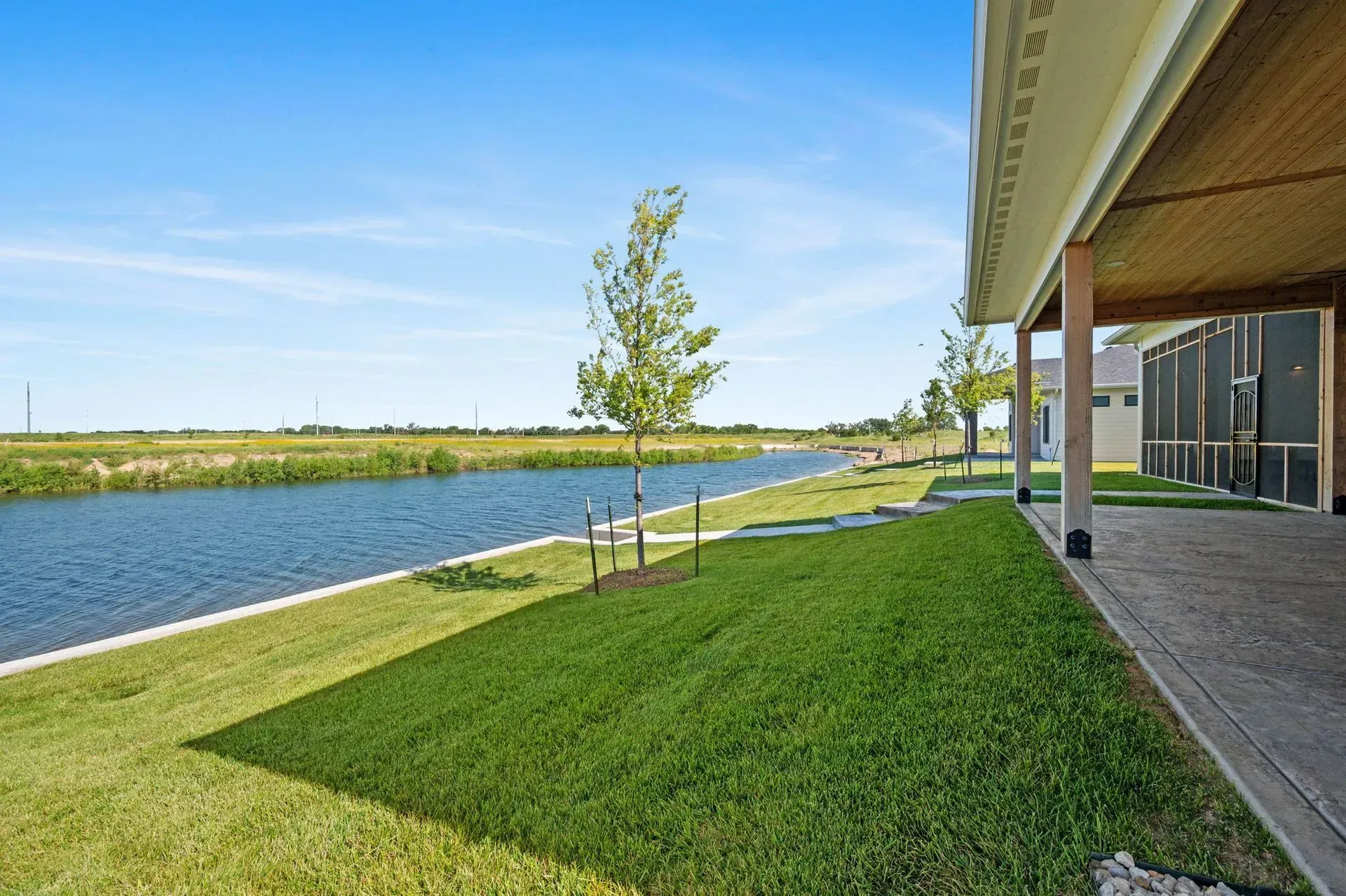 Grassy riverbank beside a canal, with a young tree, blue sky, and a house porch on the right.