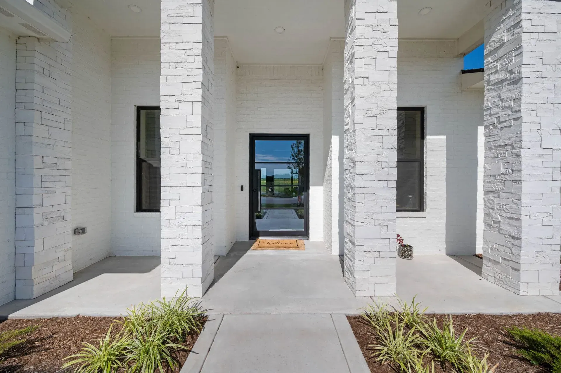 White brick home entrance with front walkway, columns, and glass door leading to a landscaped courtyard.