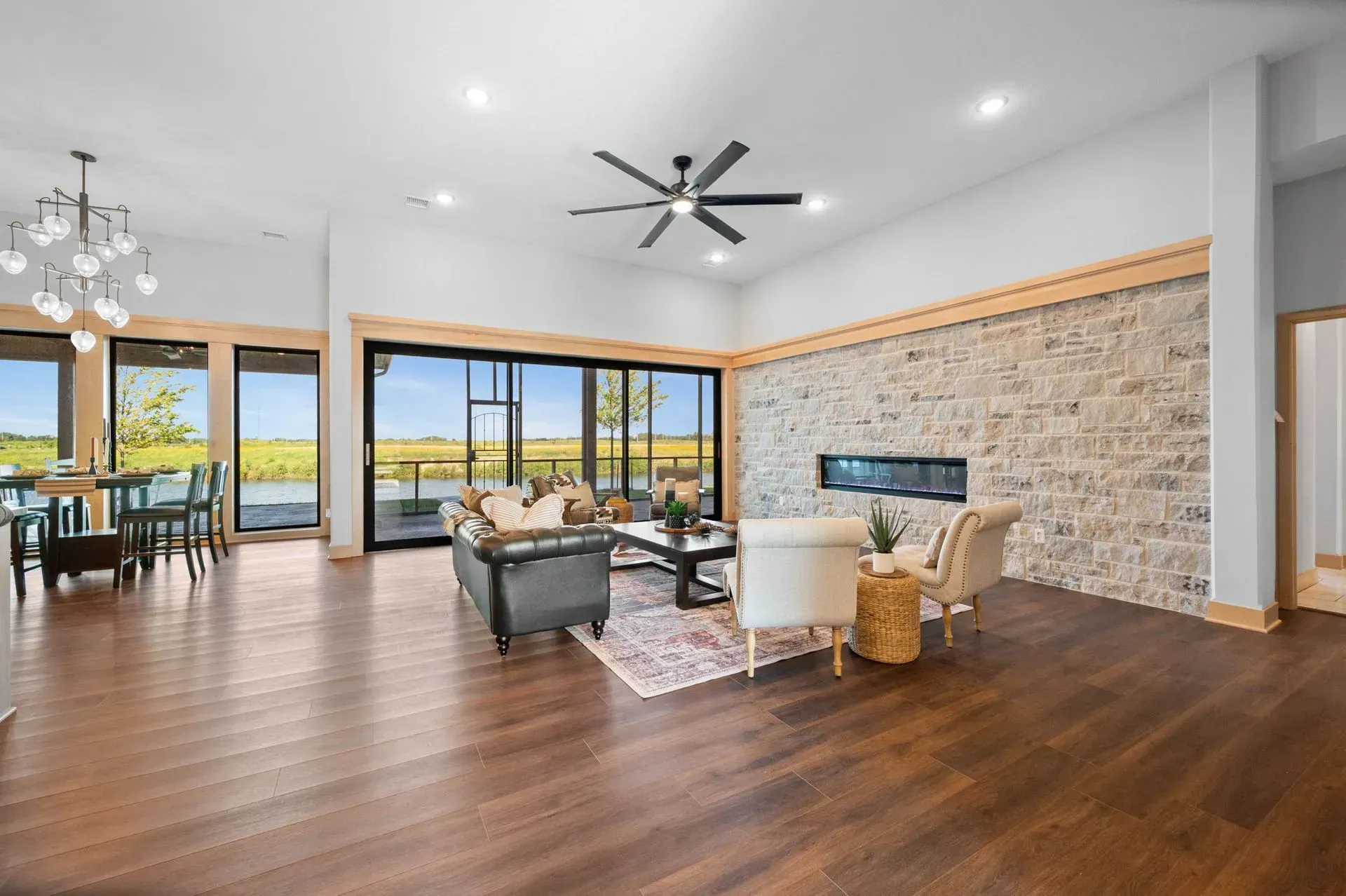 Modern open-concept living room with wood floors, stone fireplace, and large windows overlooking water.
