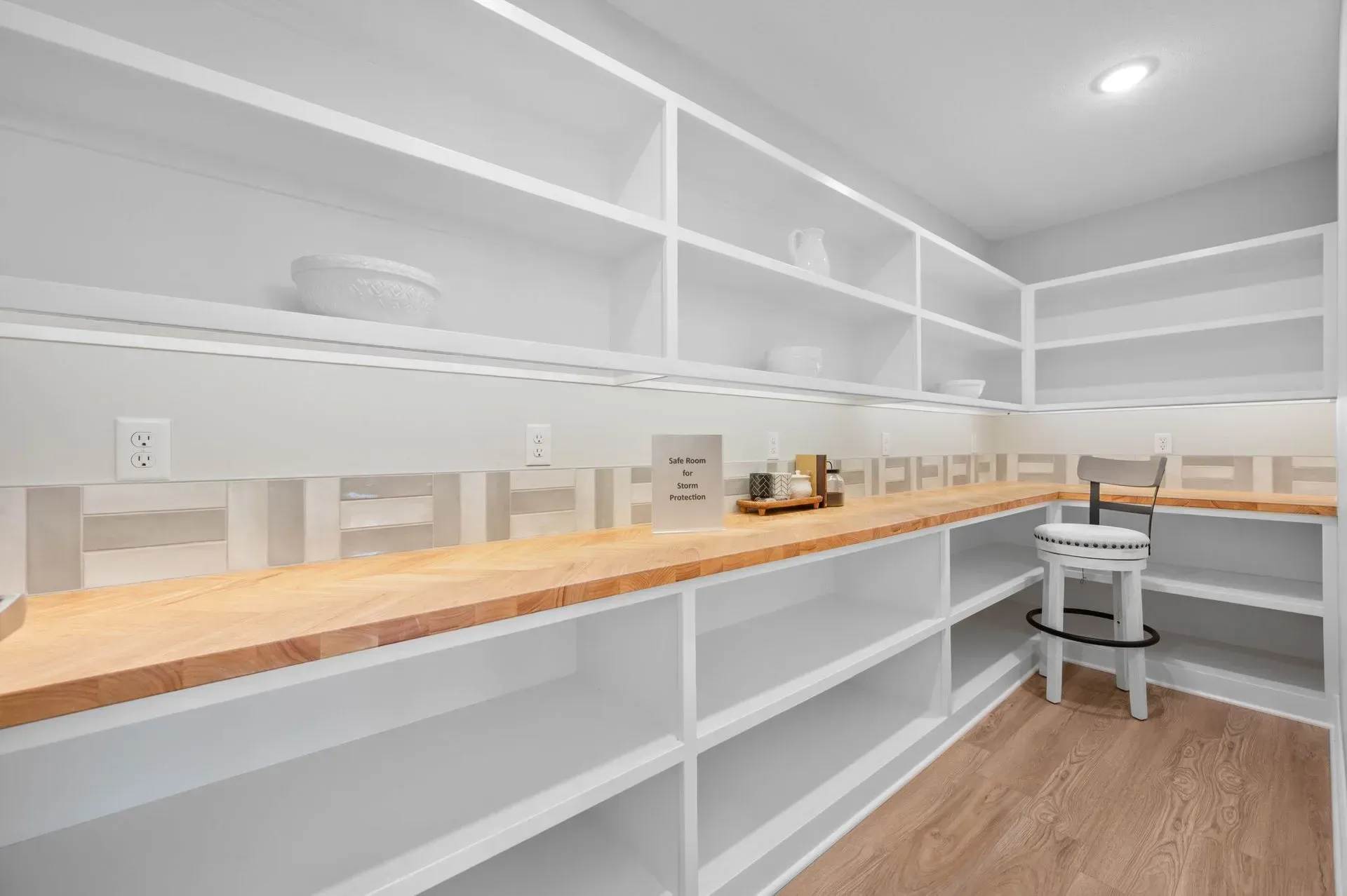 Bright white pantry with long wooden countertops, open shelving, and a stool on a hardwood floor.