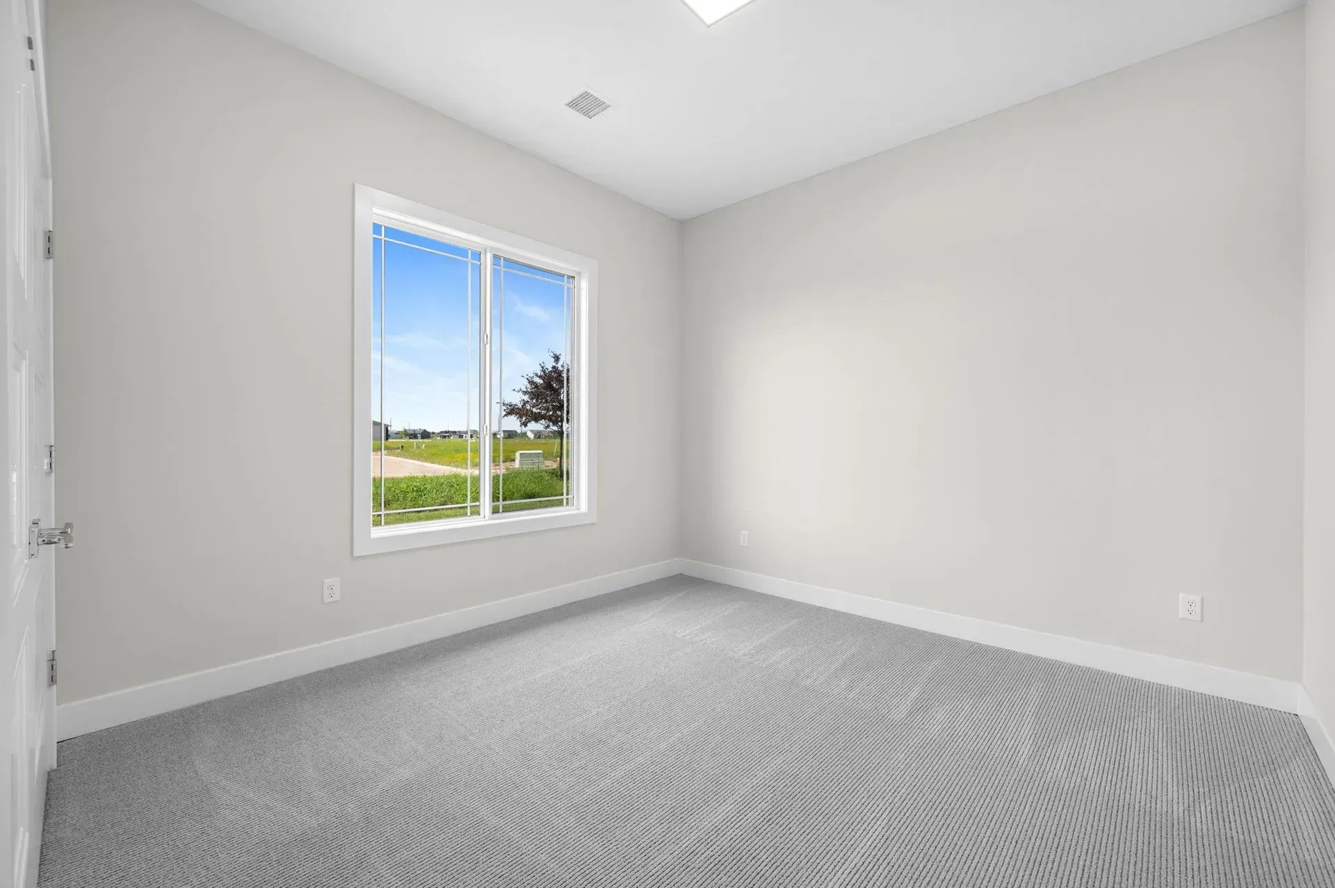 Empty white bedroom with gray carpet and a window overlooking green grass and blue sky