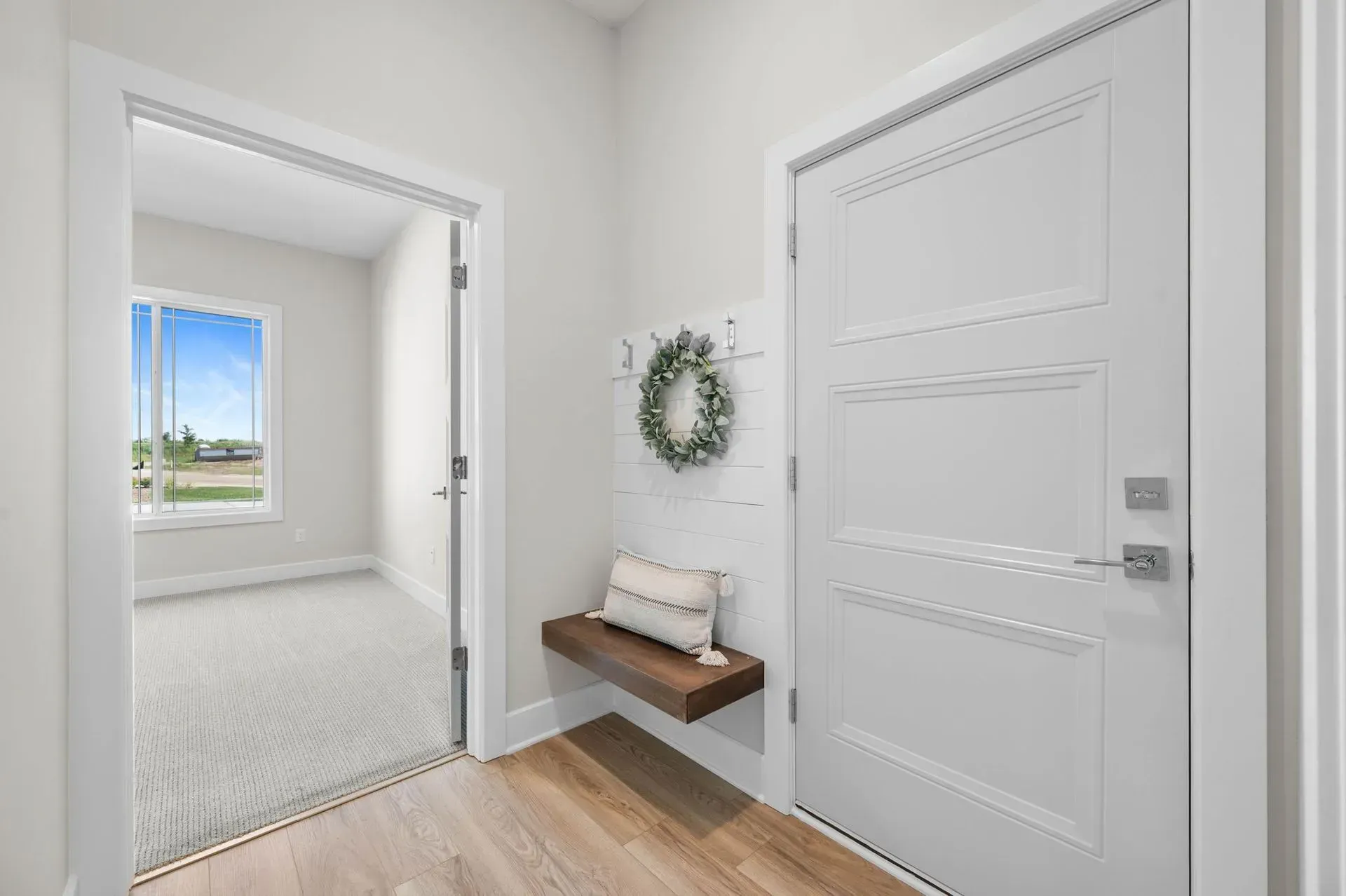 Bright entryway with white door, wall shelf, wreath, and open view into a sunlit room.