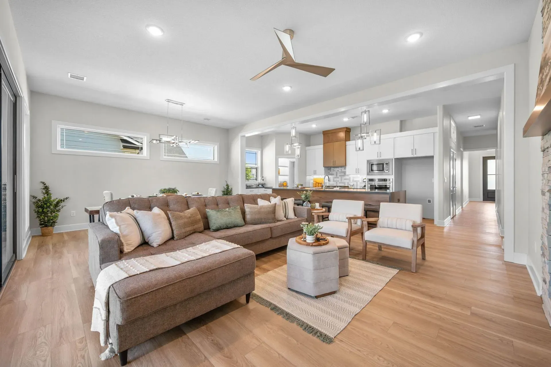 Bright open living room with sectional sofa, ottoman, wood floors, and a kitchen in the background