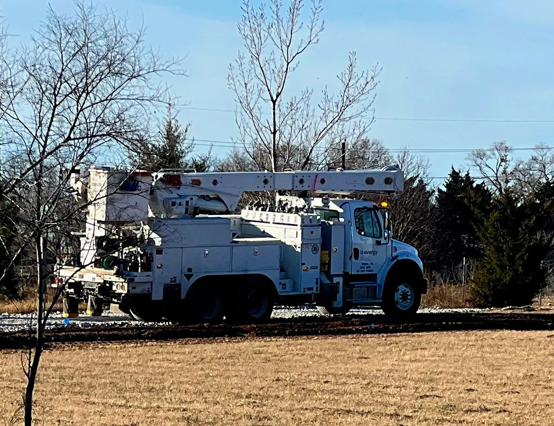 White utility truck with a boom lift parked in a field near trees under a clear sky.