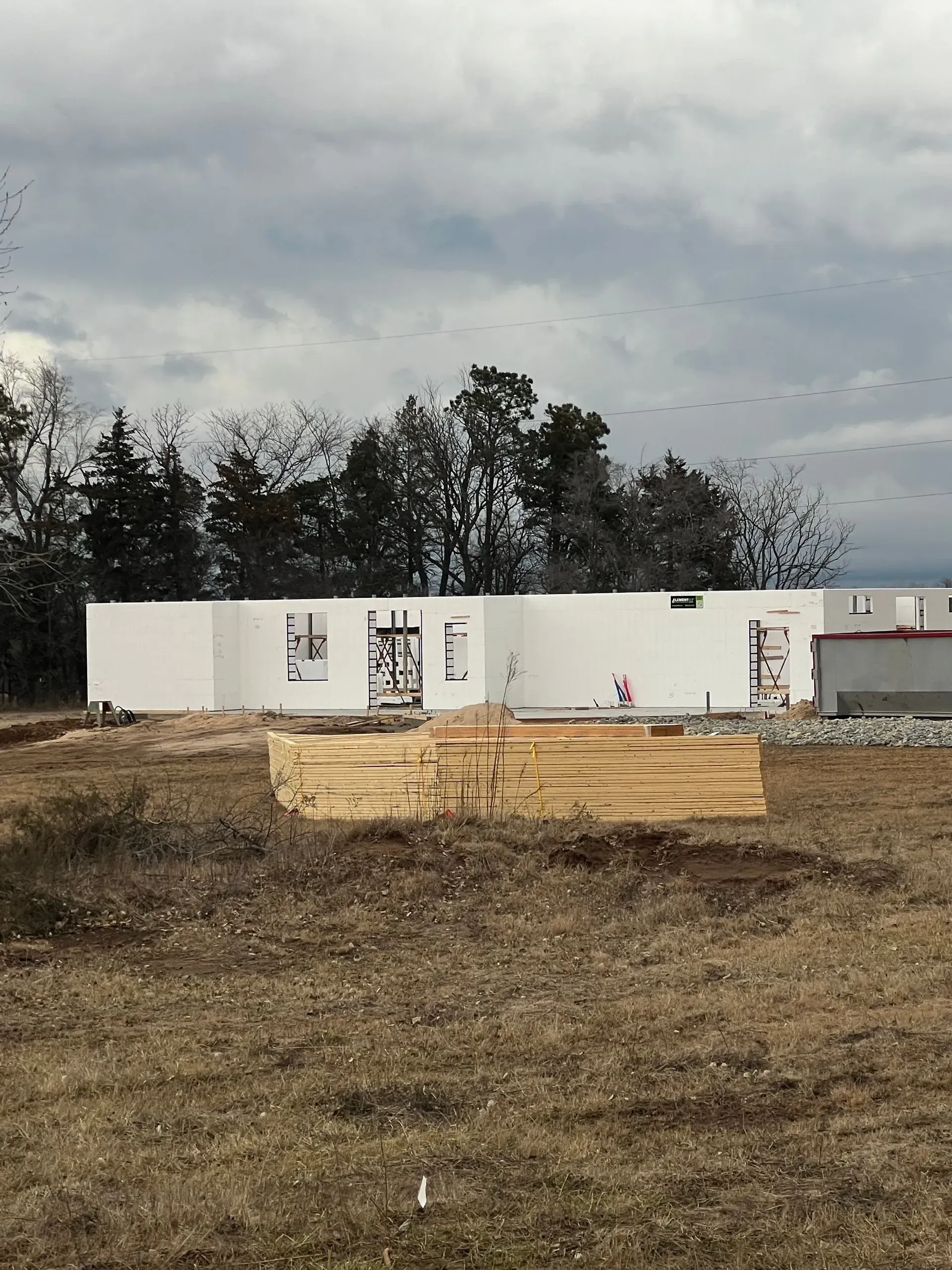 White modular building on a barren lot with stacked lumber in front under a cloudy sky