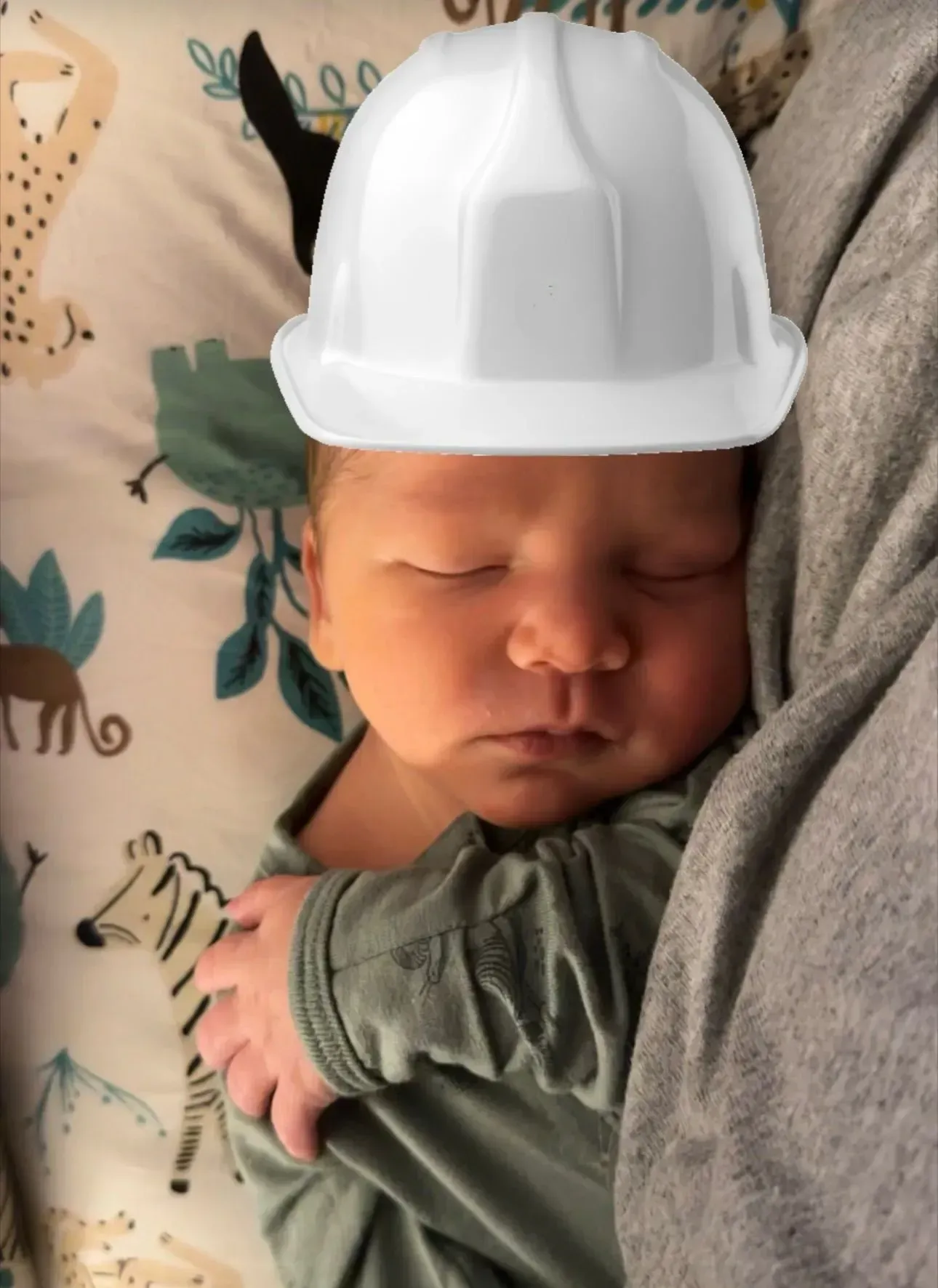 Sleeping newborn wearing a white hard hat, cradled on a patterned blanket