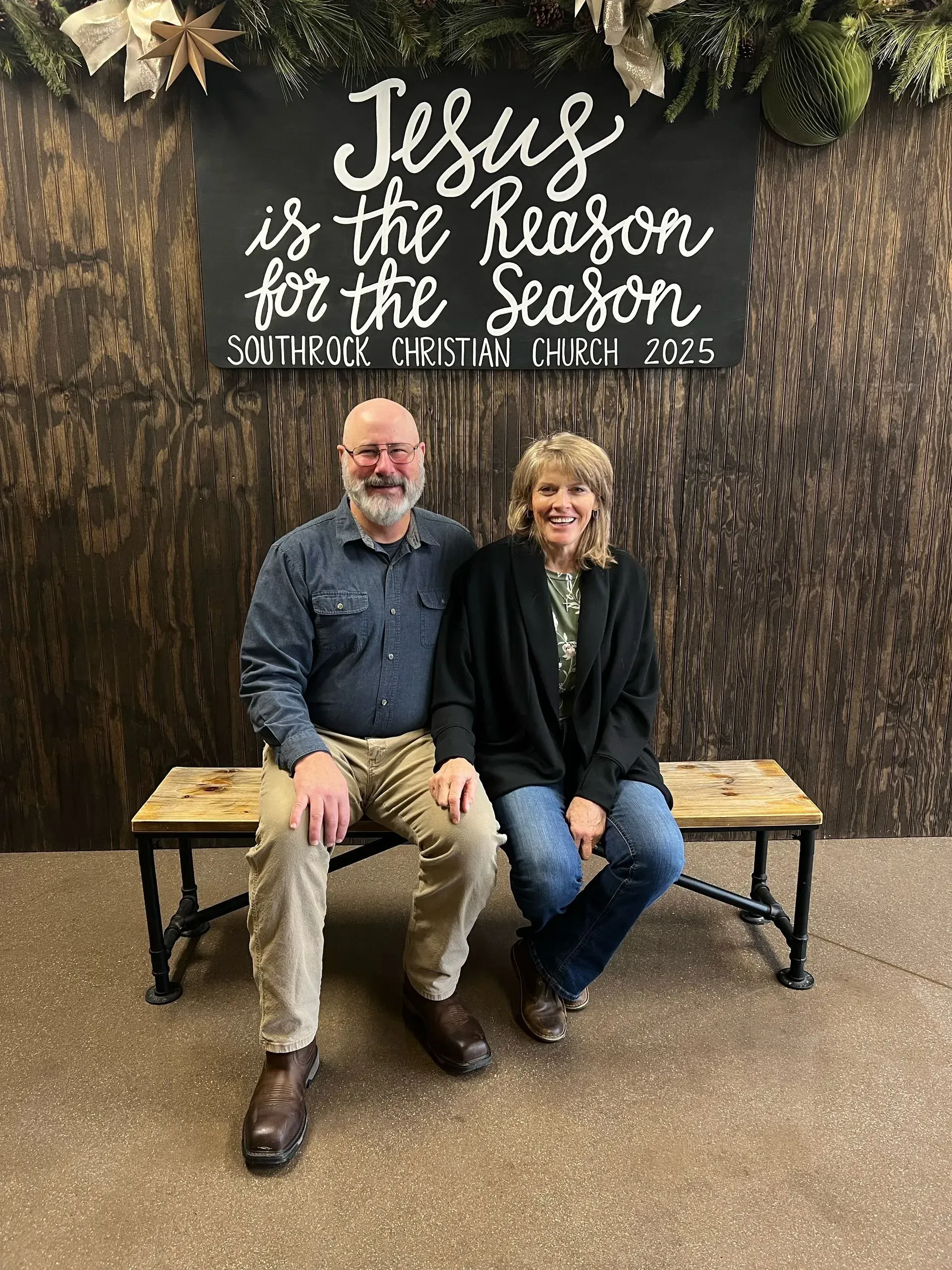 Two people sitting on a bench in front of a wall sign reading “Jesus is the Reason for the Season.”