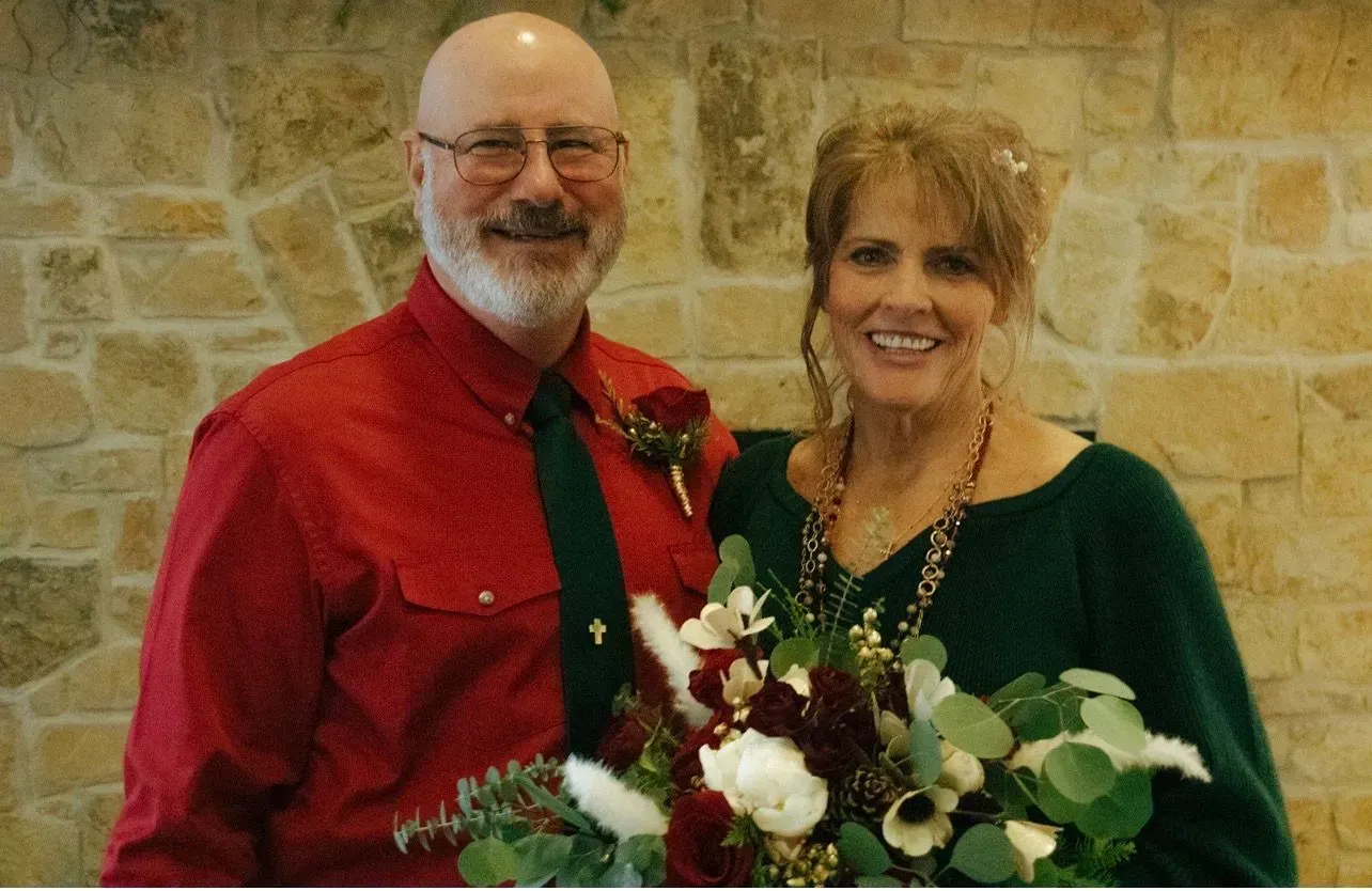 Smiling couple in festive red and green attire holding a white flower bouquet against a stone wall.