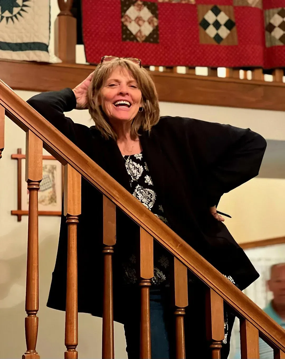 Woman smiling on a staircase with wooden banister, wearing a black jacket and jeans.