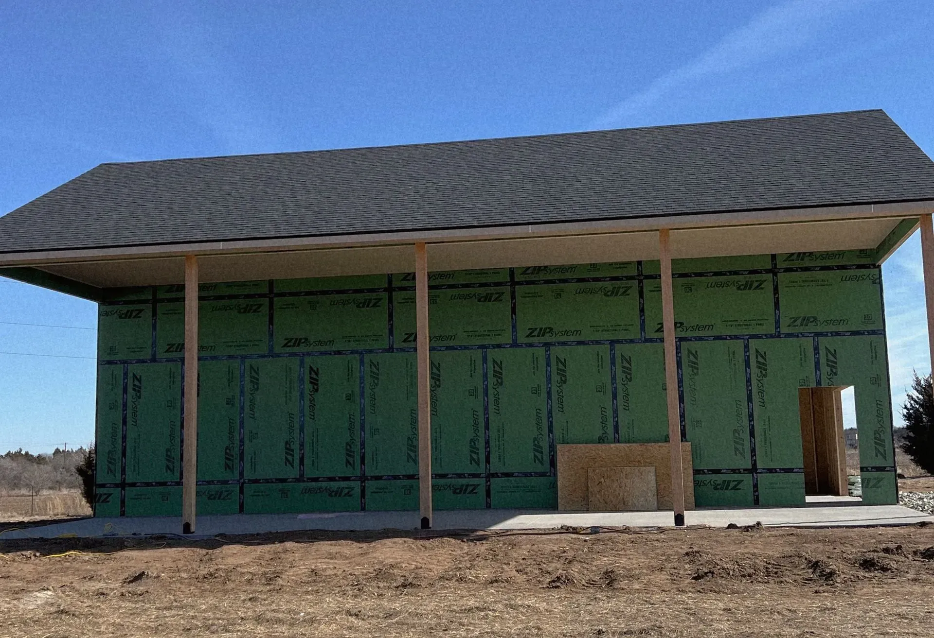 Partially built house framed in green sheathing under a gray roof on a bare dirt lot