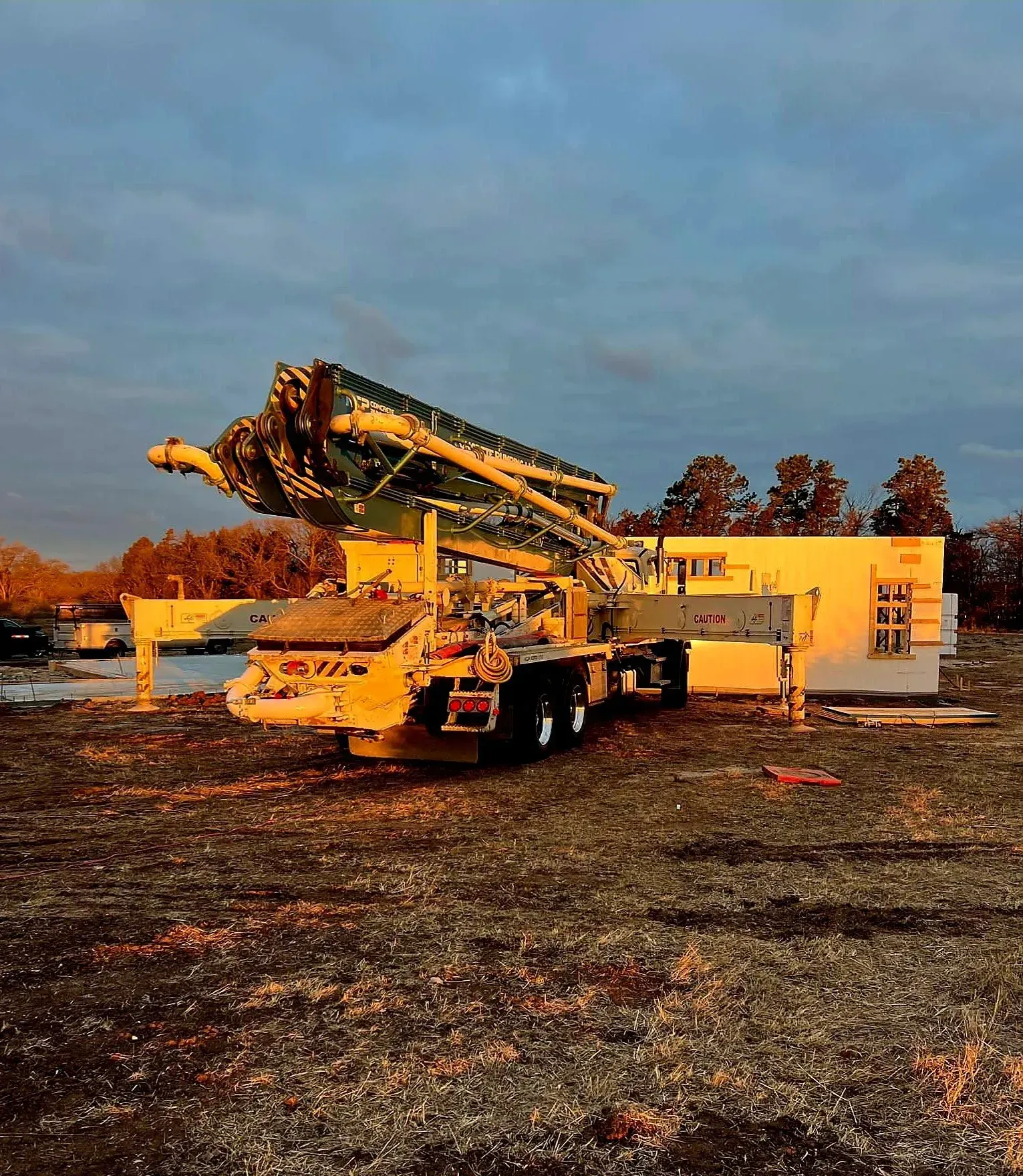 Yellow construction machine parked on a gravel lot beside a small building at sunset.