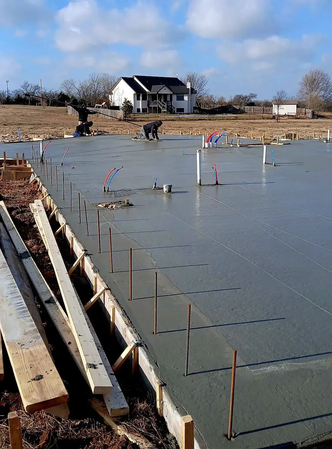 Concrete foundation slab under construction with rebar, wooden forms, and a house in the background