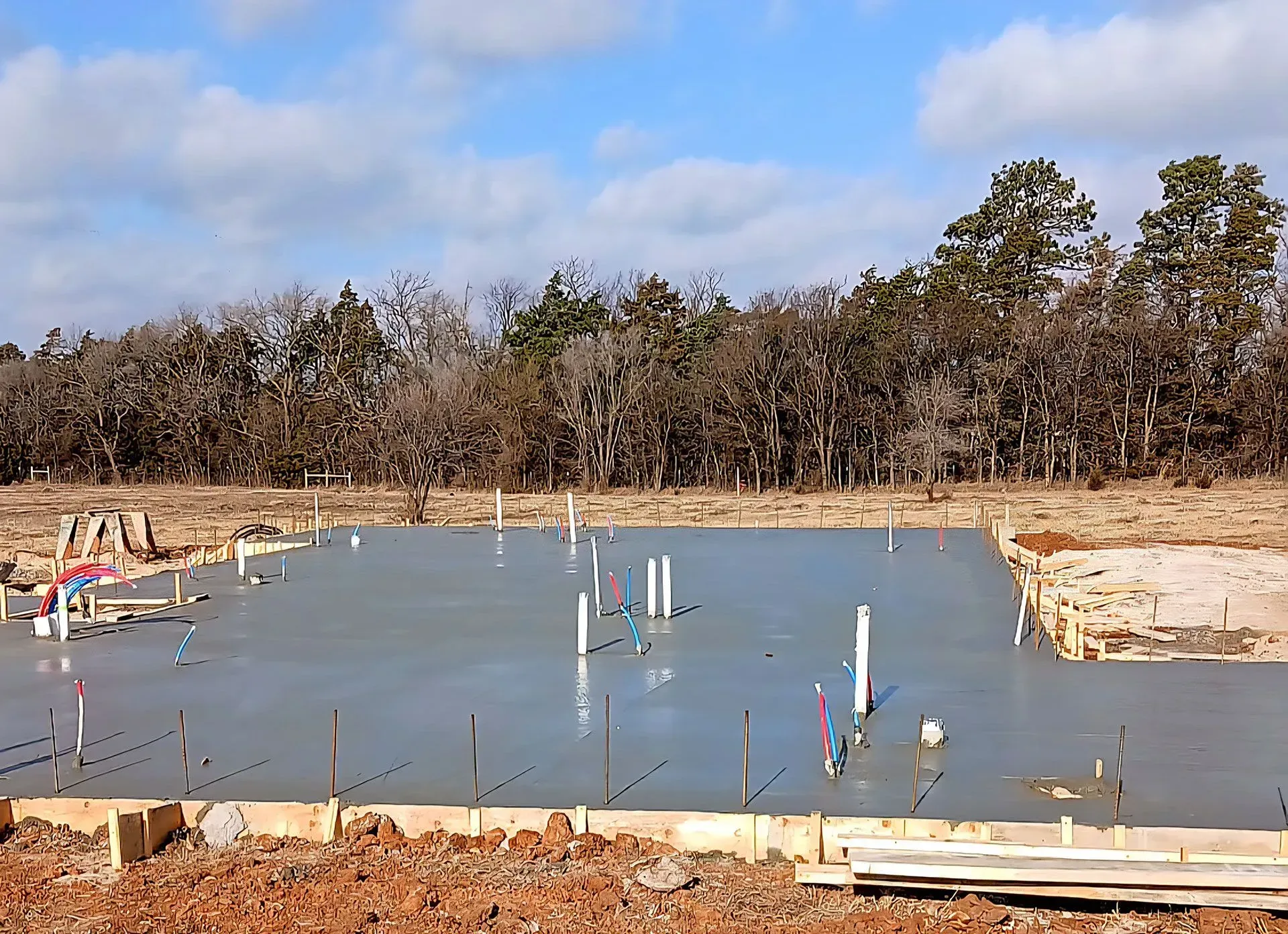 Concrete foundation slab under construction with white pipes and markers, wooded area in the background